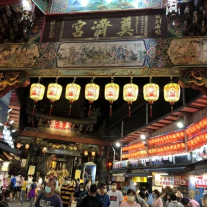 A photo of the entrance gate to the Dizang Temple in Xinzhuang Night Market, Taipei, Taiwan. The gate is intricately decorated with red and gold accents, and the entrance is surrounded by lush greenery and trees.